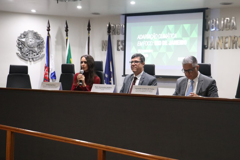 mesa retangular com uma mulher à esquerda, homem no centro e outro home à direita, com painel ao fundo com nome do evento