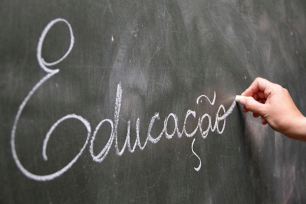 Mão segurando um giz escolar e escrevendo a palavra "Educação" em letra cursiva sobre um quadro-negro.