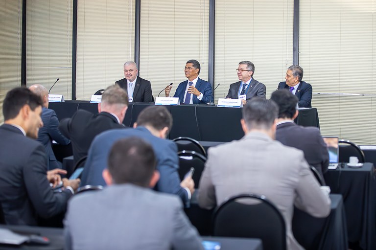 A imagem mostra uma mesa de debate com cinco homens, presumivelmente autoridades, sentados atrás de uma longa mesa preta, voltados para uma plateia. Eles estão vestidos com ternos escuros e camisas sociais. À frente de cada um, há um microfone e um crachá, embora os nomes não sejam claramente legíveis.