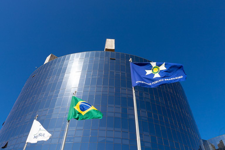 Foto do prédio A da procuradoria-geral da República. É possível ver o prédio espelhado e o céu azul sem nuvens. Em primeiro plano, as três bandeiras flamulam: a do Mercosul, a esquerda; a do Brasil, ao centro; a do Ministério Público Brasileiro, a direita