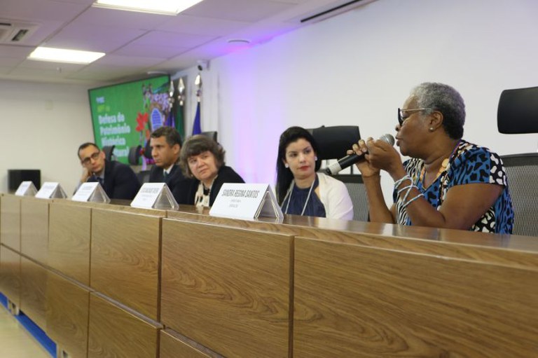 imagem de mesa larga com cinco pessoas, em primeiro plano uma mulher negra de cabelos brancos fala ao microfone.