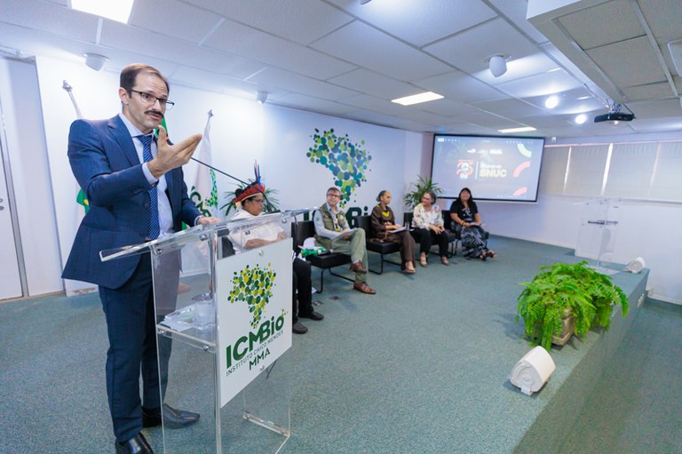 A imagem mostra o procurador da República Wilson Rocha Assis discursando em evento comemorativo dos 25 anos do SNUC, promovido pelo ICMBio. Ele fala ao microfone, em um púlpito com o logotipo do instituto, enquanto cinco pessoas — incluindo um indígena com cocar e servidores do ICMBio — o acompanham sentadas ao fundo. O ambiente é institucional, com decoração simples, plantas e uma tela de projeção exibindo a identidade visual do evento.