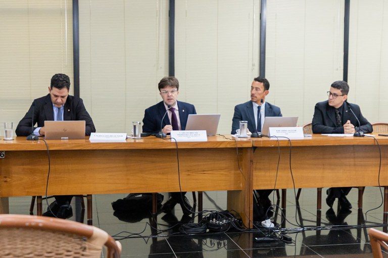 Foto da mesa de abertura do evento, com quatro homens de terno atrás de uma bancada com microfones