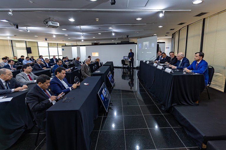 A imagem mostra uma reunião ou workshop em uma sala de conferências. Na frente da sala, há uma longa mesa com um pano preto, onde seis homens estão sentados em cadeiras de couro pretas. Todos estão vestidos com ternos e gravatas, e alguns têm microfones à sua frente. Ao fundo, uma tela de projeção exibe uma apresentação, com o nome "João Américo" visível no canto superior.