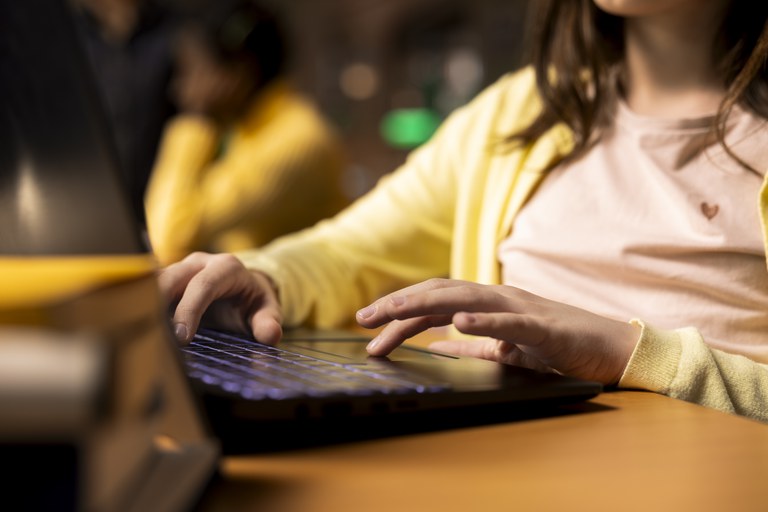 Foto de uma aluna, de pele clara que veste uma camisa rosa com coração e um casaco amarelo. Não é possível ver o rosto, mas ela tem cabeços um pouco abaixo do ombro. A estudante concentrado utiliza um computador portátil em um ambiente que sugere ser uma sala de aula ou biblioteca. O foco visual destaca as mãos no teclado.