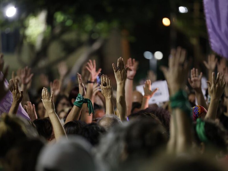 Fotografia em ambiente externo e noturno mostrando uma multidão de pessoas reunidas com os braços e as mãos levantadas para o alto. Algumas pessoas usam lenços amarrados nos pulsos, e há bandeiras de nas laterais. O foco principal está nas mãos erguidas no centro da imagem.
