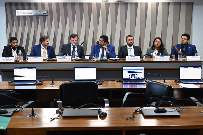 Imagem mostra seis homens e uma mulher sentados atrás de uma bancada. Em frente à bancada, duas mesas compridas, cadeiras e notebooks.