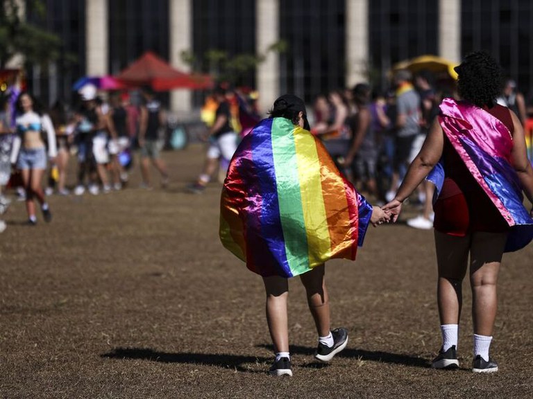 Duas pessoas caminham de costas de mãos dadas por um gramado seco durante um evento diurno ao ar livre. A pessoa à esquerda veste uma capa com as cores do arco-íris (orgulho LGBTQIA+), enquanto a da direita usa uma capa nas cores rosa, roxo e azul (orgulho bissexual). Ao fundo, várias outras pessoas aparecem desfocadas sob a luz do sol.