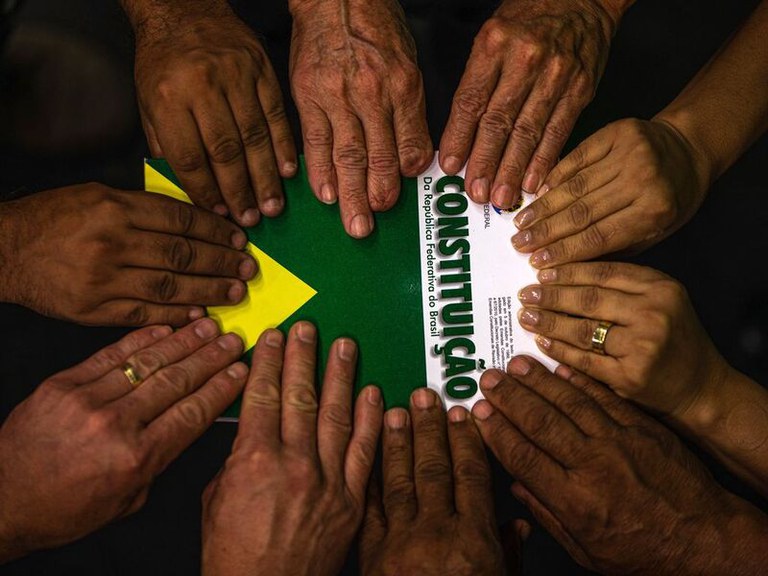 Em fundo escuro, diversas mãos com variados tons de pele estão posicionadas em círculo, repousando suavemente sobre a capa de um exemplar do livro da Constituição Federal do Brasil. A capa exibe as cores verde e amarela com o título "CONSTITUIÇÃO Da República Federativa do Brasil" escrito em letras maiúsculas de cor verde sob fundo branco.