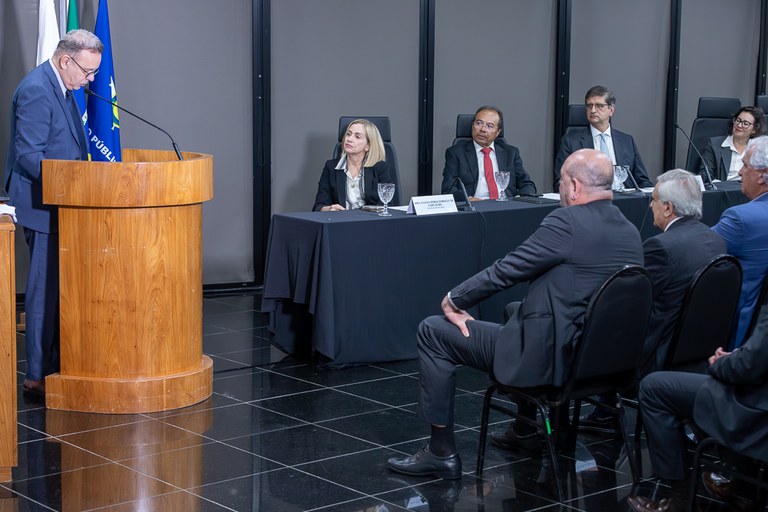 A imagem mostra um evento formal: uma pessoa fala em um púlpito diante de uma plateia, enquanto várias outras estão sentadas à mesa principal, em trajes de negócios, com bandeiras ao fundo — incluindo a do Brasil.