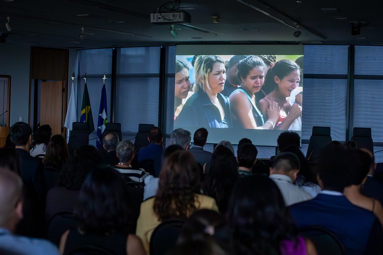 Plateia de costas em um auditório assistindo à projeção de uma foto que mostra um grupo de mulheres chorando e visivelmente emocionadas.