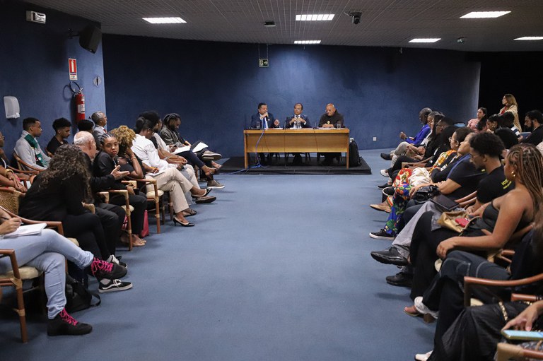 Foto mostra participantes da reunião. Em uma mesa há três pessoas