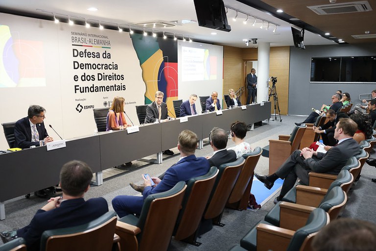 Foto mostra os participantes do evento em uma mesa com microfones a frente. Atrás há um backdrop com informações sobre o evento