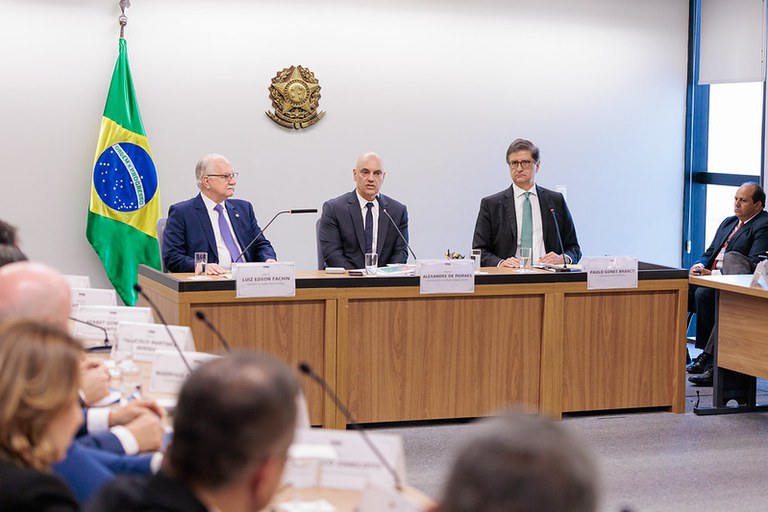 Foto de uma sala. É possível ver, da esquerda para a direita, sentados atrás de uma mesa de madeira, os ministros Edson Fachin (presidente do STF), Alexandre de Moraes (vice-presidente do STF) e Paulo Gonet Branco (procurador-geral da República). Há um brasão da República na parede e uma bandeira do Brasil ao lado do min. Fachin. Em primeiro plano, é possível ver as cabeças (de costas e desfocadas) dos procuradores-gerais de Justiça que participaram da reunião.