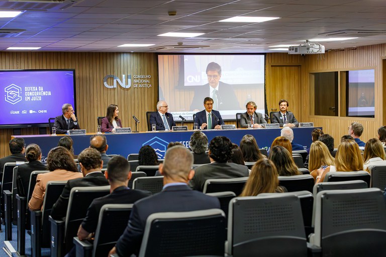 A foto mostra um auditório com público assistindo a uma mesa-redonda do Conselho Nacional de Justiça (CNJ). Seis autoridades estão sentadas na bancada sob o painel "Defesa da Concorrência em Juízo", com transmissão em um telão.