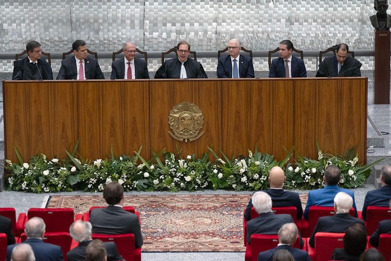 Foto da sessão de posse da nova presidência do STJ. Sete homens sentados em uma bancada de madeira com o brasão do Brasil e arranjo de flores brancas na frente. Abaixo, há um tapete e pessoas em cadeiras vermelhas assistindo à sessão solene.