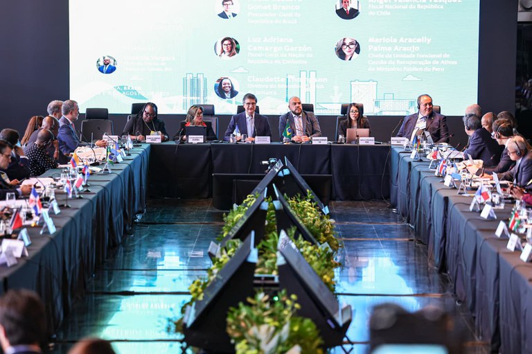 Reunião internacional em mesa em formato de U, com autoridades sentadas, pequenas bandeiras dos países sobre as mesas e um grande painel digital ao fundo exibindo fotos dos participantes.
