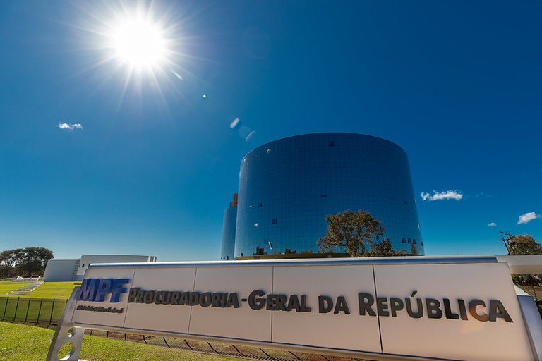 Foto dos prédios da Procuradoria-Geral da República. Em primeiro plano, há a placa de identificação do prédio com os dizeres: MPF - Ministério Público Federal (a marca do MPF na cor azul) e, ao lado, na cor preta, Procuradoria-Geral da República.
