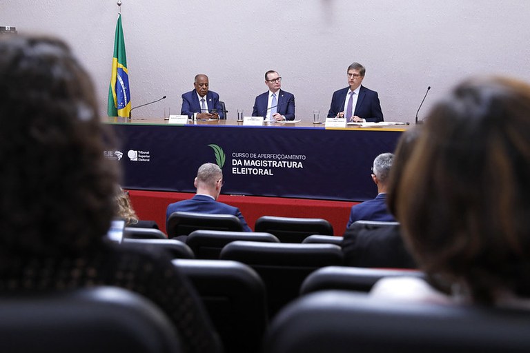 Imagem da mesa de autoridades da aula magna, com alguns participantes do curso em primeiro plano, mas de costas