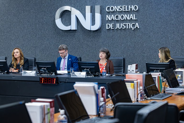 imagem de mesa retangular com três mulheres e um homem e um painel cinza no fundo escrito Conselho Nacional de Justiça