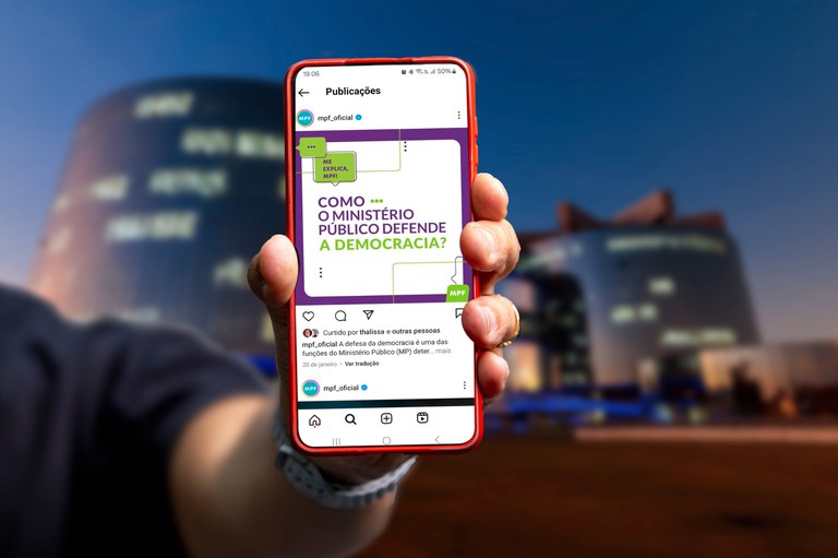 Foto de um celular com um post do "Me explica, MPF". Ao fundo, o prédio da Procuradoria-Geral da República.