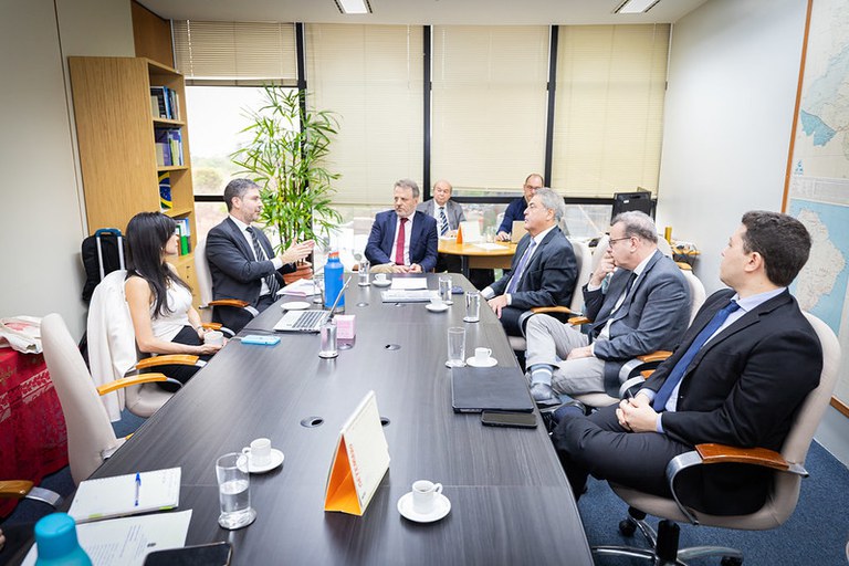 Foto da reunião de trabalho. Há oito pessoas, sete homens e uma mulher, sentados ao redor de uma mesa longa e escura. Eles vestem trajes formais. Um dos homens, posicionado ao fundo, no canto esquerdo da mesa, está gesticulando enquanto fala com os demais. Sobre a mesa há garrafas de água, copos e notebooks. A sala tem estantes com livros e pastas e uma grande janela com persianas.