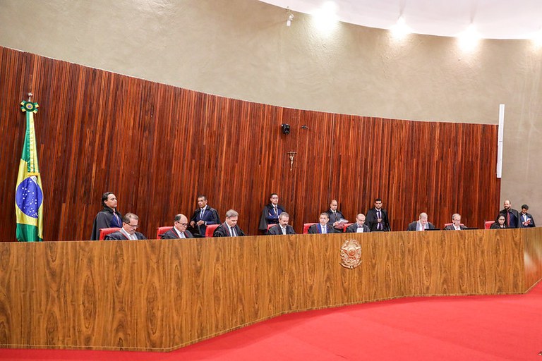 Sessão plenária no Tribunal Superior Eleitoral (TSE). Ministros e autoridades vestindo togas pretas aparecem sentados enfileirados atrás de uma longa bancada de madeira com o brasão da República ao centro. O ambiente possui um tapete vermelho no chão, uma parede de fundo revestida por ripas verticais de madeira com um crucifixo fixado e a bandeira do Brasil posicionada à esquerda.