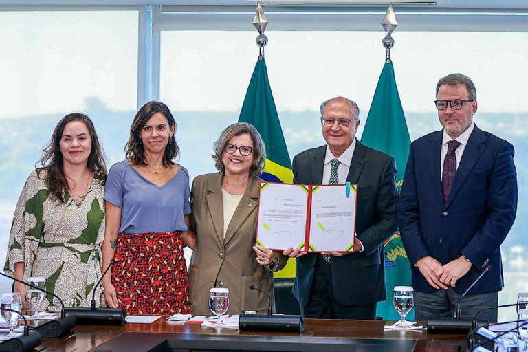 Foto mostra o vice-presidente, Geraldo Alckmin, ao lado do vice-procurador-geral da República, Hindenburgo Chateaubriand e de outas três mulheres, enquanto segura um documento oficial recém-assinado, com a bandeira do Brasil ao fundo.