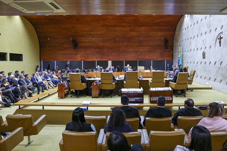 A imagem, tirada de lado, mostra o interior do Plenário do STF, com a corte em sessão. Além dos 11 ministros, o procurador-geral da República ocupa a cadeira ao lado do presidente do STF. Estão sentados em uma bancada elevada, de madeira escura, no centro da sala. No nível inferior, estão os advogados (lado esquerdo da foto). De costas para a câmera, o público, com alguns ocupantes da sala. Todos estão em cadeiras de couro marrom, alinhadas em filas. A sala, com iluminação amarelada e paredes de madeira.