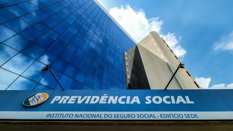 Foto de parte da fachada do prédio do INSS. Ele está fotografado de baixo para cima. O prédio é espelhado e é possível ver nuvens no céu. Em primeiro plano, está escrito na marquise "Previdência Social - Instituto Nacional do Seguro Social - Edifício Sede"