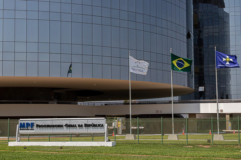 Foto do prédio da PGR com destaque para o letreiro da Procuradoria-Geral da República e para as bandeiras do Mercosul, do Brasil e do Ministério Público Brasileiro (da esquerda para a direita).