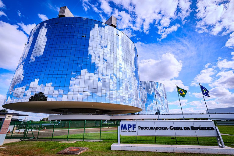 Foto dos prédios da Procuradoria-Geral da República. A frente, a placa do prédio com a logo do MPF, em azul, e o nome Procuradoria-Geral da República, em preto. É possível ver as bandeiras do Mercosul, do Brasil e do Ministério Público O céu está azul e há nuvens.
