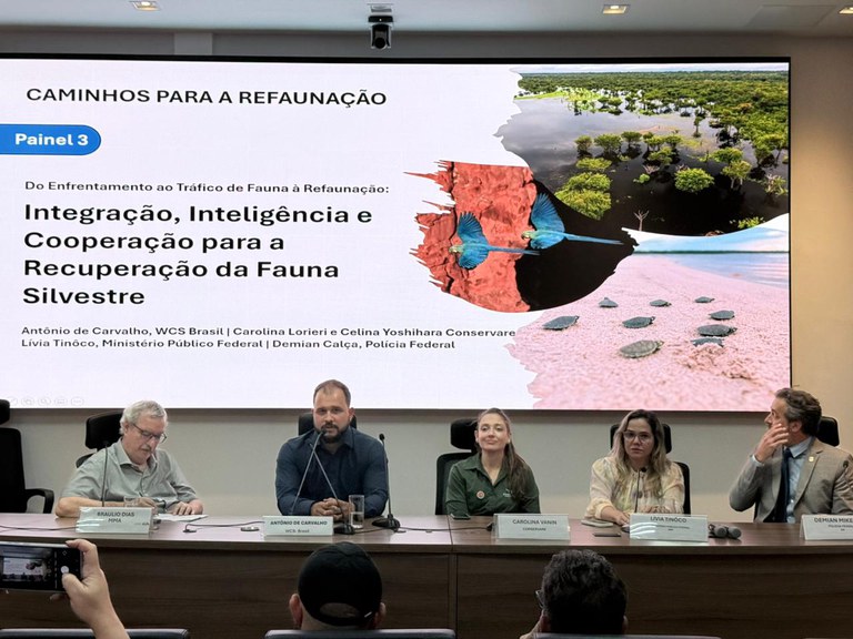 mesa longa com cinco pessoas e ao fundo um painel com o título do evento