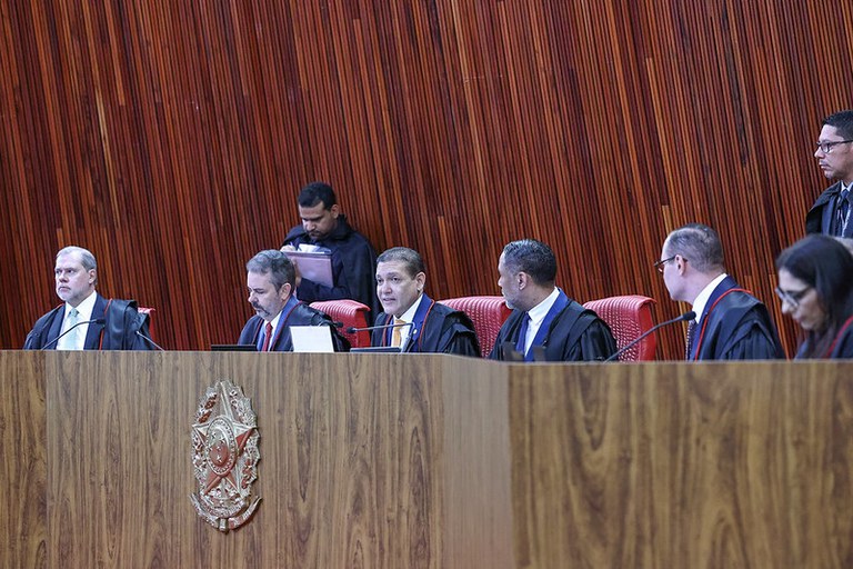 Imagem mostra a sessão de julgamento no plenário do Tribunal Superior Eleitoral (TSE) do dia 28/5. Os ministros da Corte e o Vice-PGE estão sentados atrás de uma bancada de madeira, vestindo as togas pretas. Atrás da bancada, de pé, há assessores vestindo uma capa preta e manuseando alguns papéis.