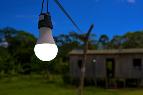 Fotografia de uma lâmpada acesa, em primeiro plano, pendurada por um fio elétrico, com uma casa ao fundo, desfocada, com o céu escurecendo