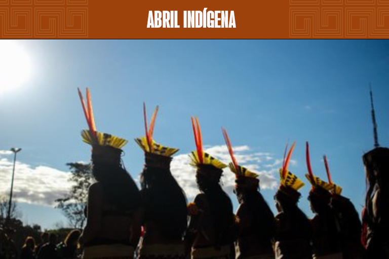 Fotografia ilustrativa mostra mulheres indígenas de costas na contraluz, com seus cocares e adereços tradicionais, em contraste com o céu azul