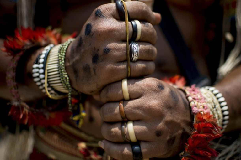 Plano detalhado das mãos de uma pessoa indígena com pinturas corporais escuras, usando diversos anéis e pulseiras artesanais de contas e penas, enquanto segura firme um bastão de madeira.