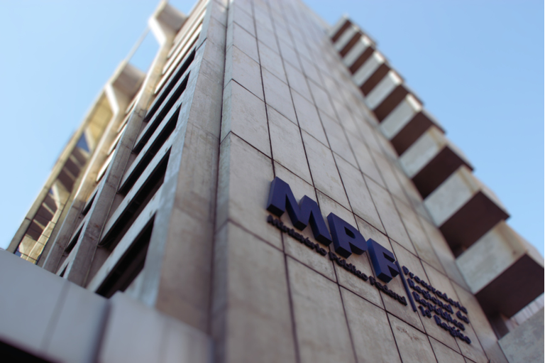 Fotografia colorida em ângulo baixo (contra-plongée) do edifício da Procuradoria Regional da República da 1ª Região. A imagem destaca a fachada de concreto em tons de bege claro, com janelas verticais e detalhes arquitetônicos que dão sensação de altura. No centro, fixada na parede, vê-se a sigla "MPF" em letras azul-escuras em relevo, acompanhada do texto "Ministério Público Federal" e a identificação da unidade. O céu ao fundo é azul claro e sem nuvens, com a luz solar iluminando a estrutura lateralmente.