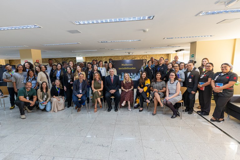 Participantes da abertura da exposição posam para foto. Eles estão próximos aos painéis da exposição, sendo os procuradores sentados em cadeiras ao centro. Os demais estão em pé.