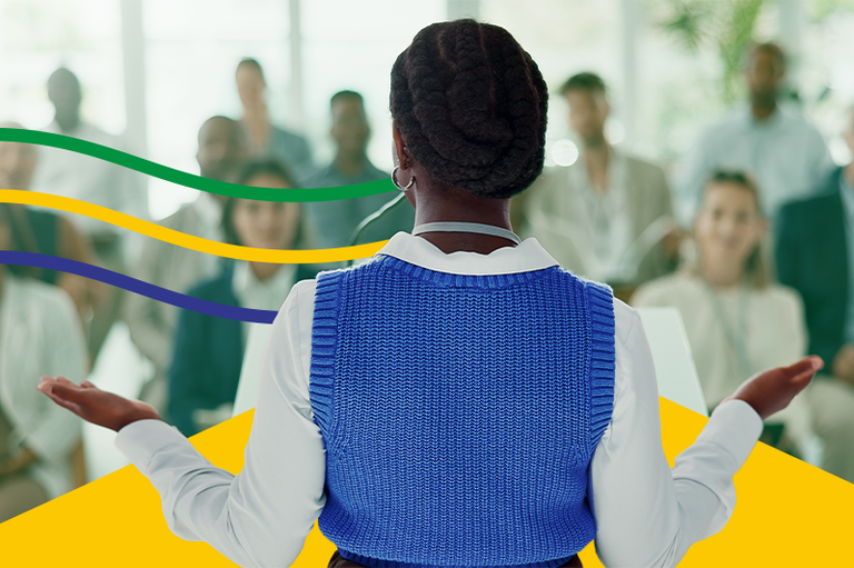 Uma mulher negra é vista de costas, no centro do primeiro plano, falando para uma plateia desfocada durante uma apresentação ou discurso. Ela está com as mãos abertas, usando um colete de tricô azul sobre uma blusa de gola branca e um penteado trançado. Ao fundo, uma plateia diversificada sentada a assiste atentamente. Elementos gráficos com as cores da bandeira do Brasil emolduram a imagem, com três linhas onduladas nas cores verde, amarela e azul à esquerda e detalhes em amarelo vibrante nos cantos inferiores.