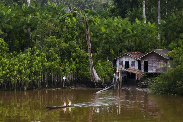 Duas pessoas em uma pequena canoa navegando por um rio, com casas de madeira ribeirinhas e densa vegetação tropical ao fundo.