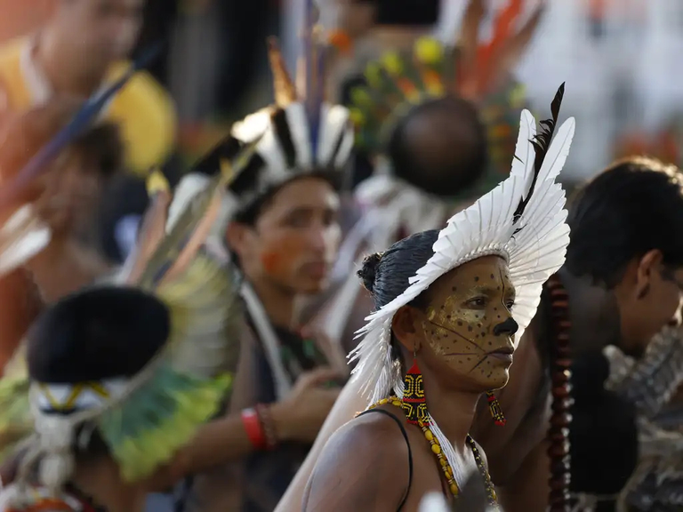 Fotografia colorida em close-up de perfil de uma mulher indígena em primeiro plano, com pintura facial amarela e preta que imita manchas de onça e um cocar branco grande feito de penas. Ela tem o cabelo escuro preso e usa um top ou alça preta com colares de miçangas vermelhas e amarelas. Ao fundo, fora de foco, outros participantes com adereços e pinturas faciais tradicionais parecem estar em uma concentração ou evento. A iluminação é forte, sugerindo um ambiente externo de dia.