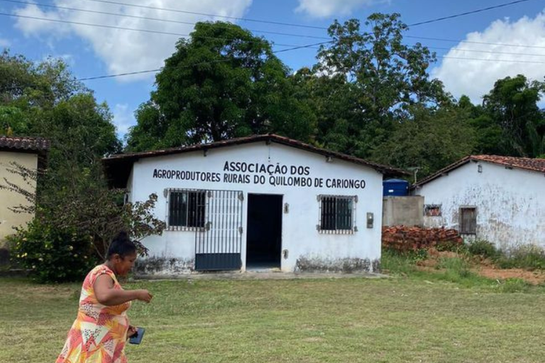 Fachada de um edifício branco com a inscrição «Associação dos Agroprodutores Rurais do Quilombo de Cariongo». Uma mulher com vestido estampado caminha em primeiro plano num dia de sol.