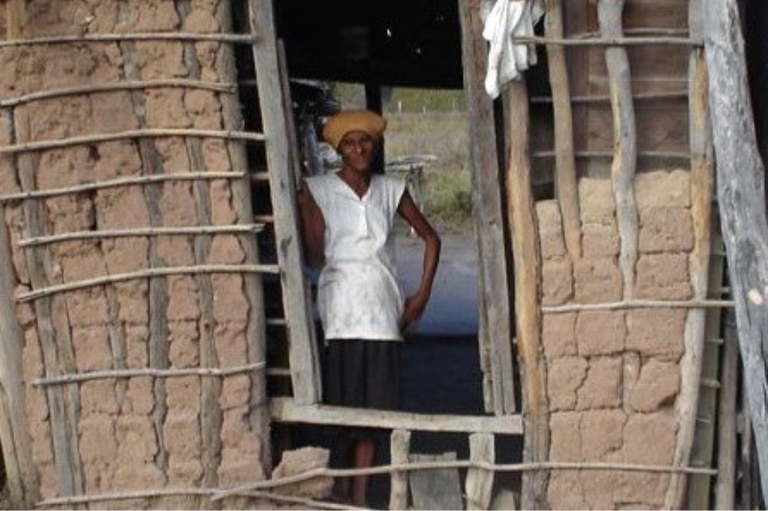 Mulher negra idosa em pé no vão de uma porta de uma casa de pau a pique (taipa). Ela veste uma blusa branca, saia escura e um turbante amarelado. As paredes da construção mostram a estrutura de madeira e o barro seco com rachaduras naturais.