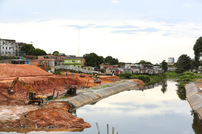 Foto mostra obras em margem de igarapé. Ao fundo, vê-se casas construídas bem perto do igarapé. Na margem esquerda, há retroscavadeiras