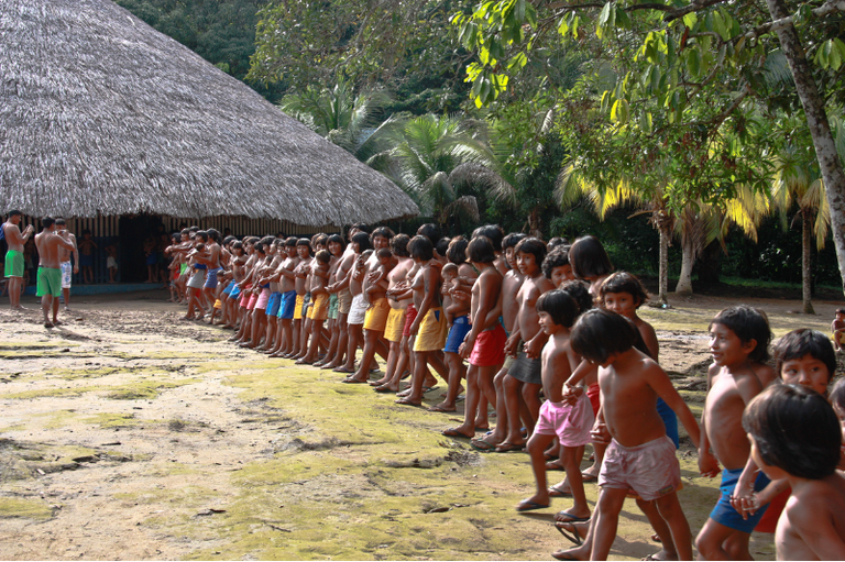 Um grupo de crianças e adolescentes indígenas Waimiri Atroari está em uma fila longa em frente a uma oca ou casa comunitária com telhado de palha. A maioria veste apenas shorts coloridos, e o ambiente é de terra batida sob a luz solar, com vegetação densa de floresta ao redor.