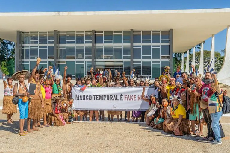 Foto de indígenas em frente ao STF com uma faixa onde está escrito Marco Temporal é Fake News