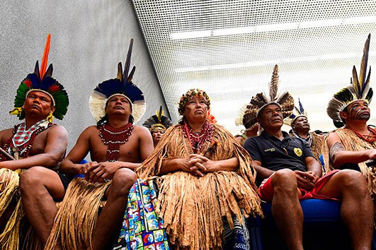 A imagem mostra um grupo de indígenas sentadas em um ambiente fechado. Elas vestem trajes tradicionais, com cocares coloridos e adornos de penas, colares e saias de palha. Algumas pessoas também usam roupas comuns, como camisetas.