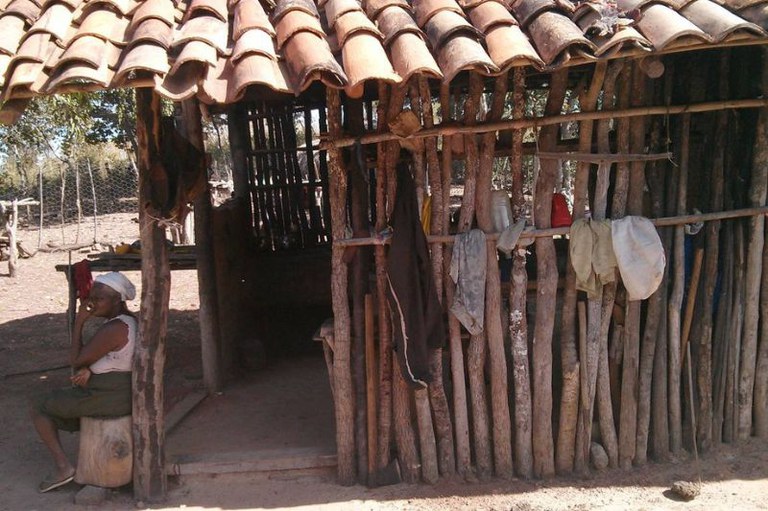 Foto mostra um barraco com estacas de madeira, algumas roupas penduradas e uma senhora sentada ao lado