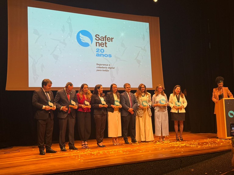 Foto de um palco, em que oito procuradores recebem uma placa cada de premiação. Ao fundo, a projeção da Safernet.